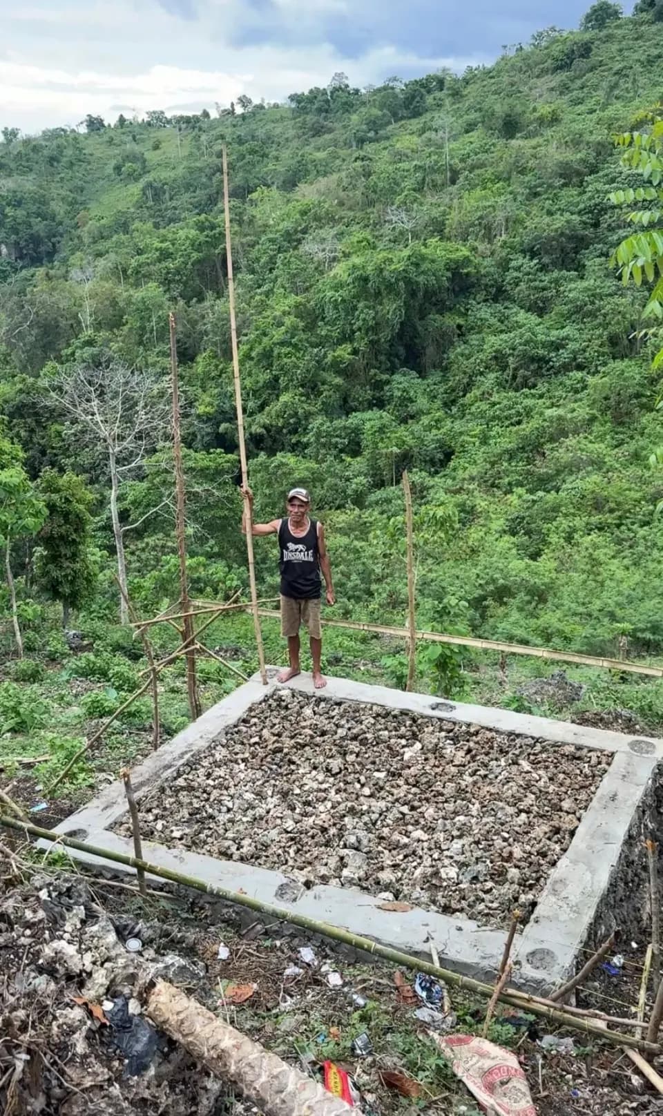Seorang warga berdiri di atas fondasi beton tangki yang baru dibuat, dengan lereng bukit berhutan di belakangnya.