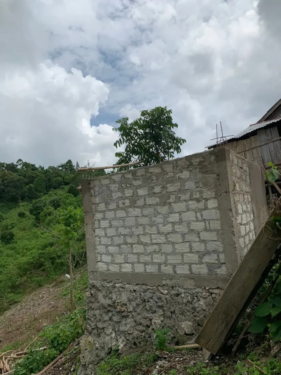Dinding batako tangki yang sebagian sudah diplester, berdiri di atas dasar batu di bawah langit berawan.