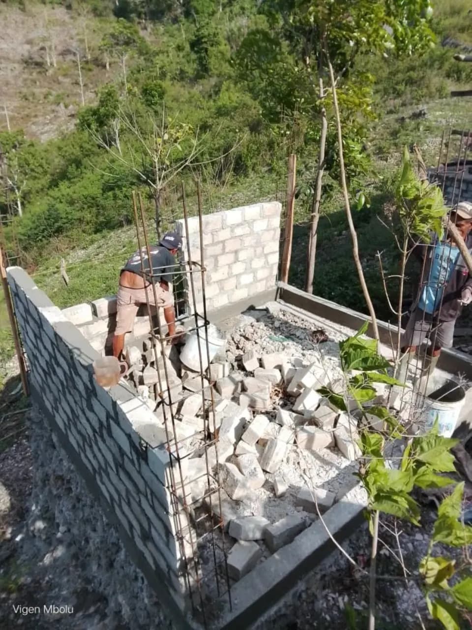 Dua warga memasang dinding batako tangki di antara tumpukan batu dan besi tulangan.