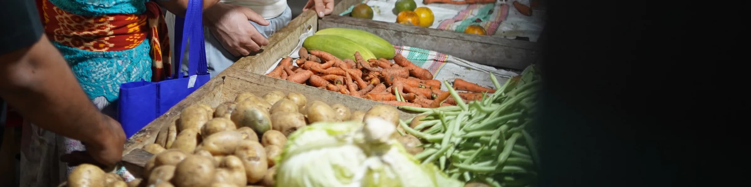 Peti kentang, wortel, buncis, dan kubis di sebuah lapak pasar tertutup di Sumba, dengan tangan penjual di belakangnya.